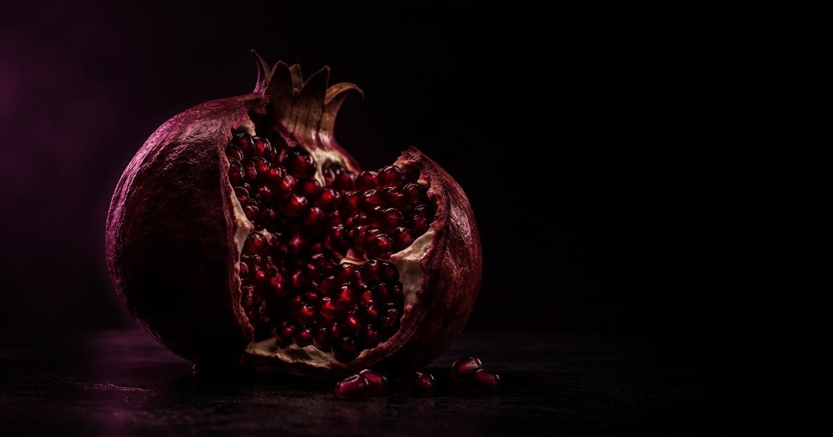 A pomegranate split open against a near-black background, lit in deep mulberry